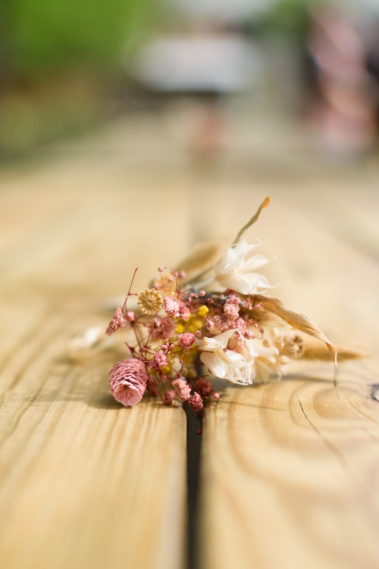 Un petit bouquet de fleurs roses et blanches séchées repose sur une surface en bois. L'arrière-plan est flou, attirant l'attention sur les textures et les couleurs délicates des fleurs reposant au centre de l'image.