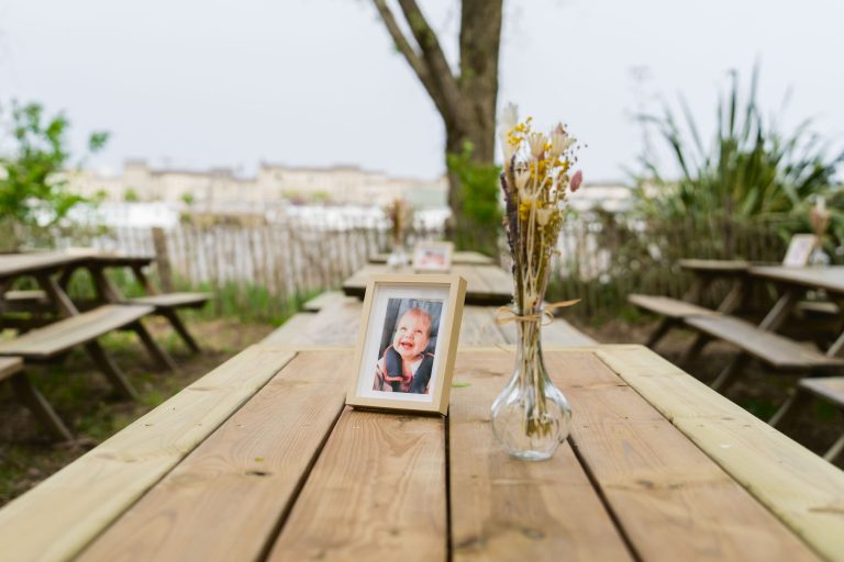 Une table de pique-nique en bois à l'extérieur contient une photo encadrée d'un enfant souriant et un vase en verre transparent avec des fleurs séchées. D'autres tables de pique-nique et de la verdure se trouvent à l'arrière-plan, avec des arbres et des bâtiments flous visibles au loin.