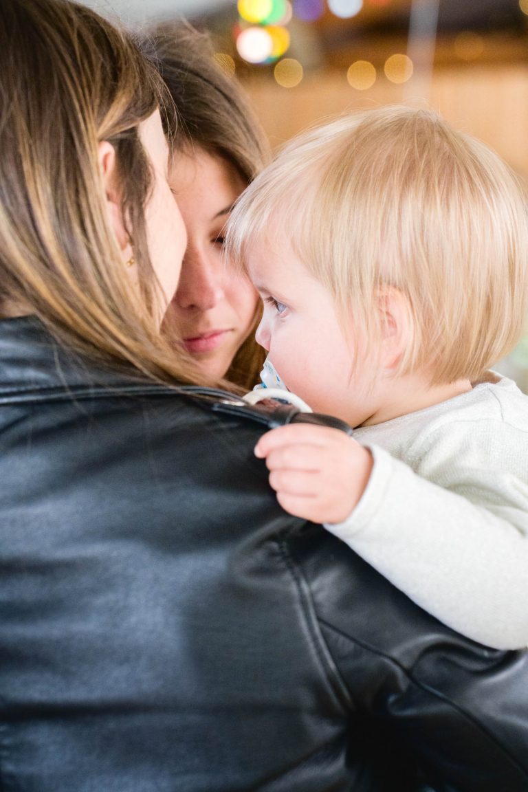 Un bambin blond avec une tétine repose sur l'épaule d'un adulte vêtu d'une veste en cuir noir, tandis qu'une autre personne aux longs cheveux bruns regarde l'enfant, les deux adultes étant partiellement visibles et proches l'un de l'autre, ce qui crée un moment d'intimité et d'attention.
