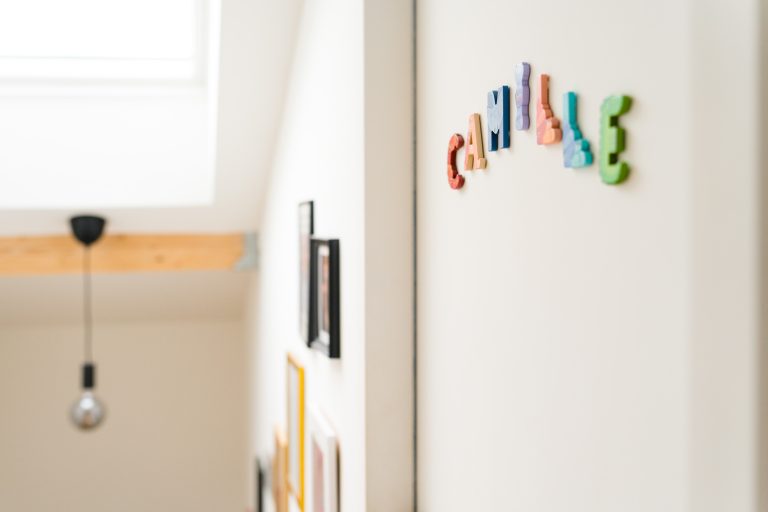 Des lettres magnétiques colorées épelant "SMILE" sont fixées sur un mur blanc dans un espace lumineux et moderne. Des photos encadrées sont accrochées au mur adjacent. Une ampoule nue avec une fixation noire et une poutre en bois est visible près d'une grande fenêtre au-dessus.
