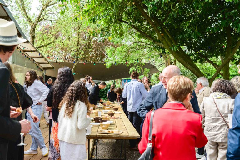Un groupe de personnes se réunit en plein air, sous des arbres feuillus, autour d'une longue table en bois garnie de nourriture et de boissons. Certains restent debout et discutent, tandis que d'autres se servent eux-mêmes. La scène est animée et ensoleillée, et l'on peut voir divers vêtements décontractés et semi-formels.