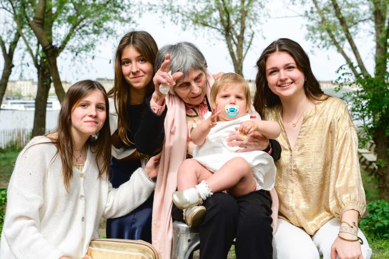 Quatre jeunes femmes et une femme âgée sont assises à l'extérieur et sourient à la caméra. La femme âgée tient un bébé avec une tétine sur ses genoux et fait un geste ludique. Les arbres et l'eau sont à l'arrière-plan, et tout le monde a l'air heureux et détendu.