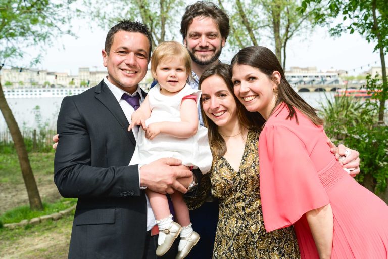 Cinq personnes se tiennent près les unes des autres en plein air et sourient à l'appareil photo. Un homme en costume tient un enfant en bas âge vêtu d'une robe blanche. Deux femmes, l'une en robe à motifs et l'autre en robe corail, se tiennent à côté d'eux. Des arbres verts et une rivière apparaissent à l'arrière-plan.