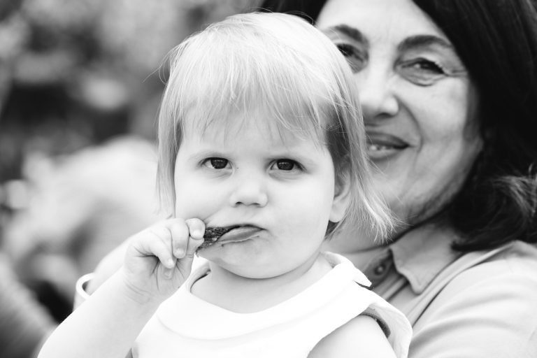 Un jeune enfant aux cheveux clairs regarde l'appareil photo tout en portant de la nourriture à sa bouche. Une femme plus âgée aux cheveux foncés sourit à l'arrière-plan, partiellement floue. L'image est en noir et blanc, créant une atmosphère douce et candide.