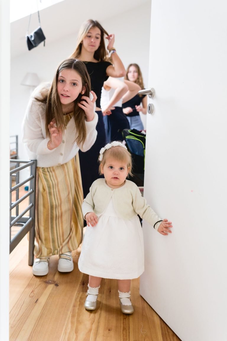 Un enfant en bas âge vêtu d'une robe blanche se tient près d'une porte ouverte sur un sol en bois, tandis qu'une jeune fille vêtue d'une jupe à rayures s'accroupit à ses côtés. Trois autres filles se tiennent derrière elles dans une pièce moderne et lumineuse, et regardent toutes vers la caméra.