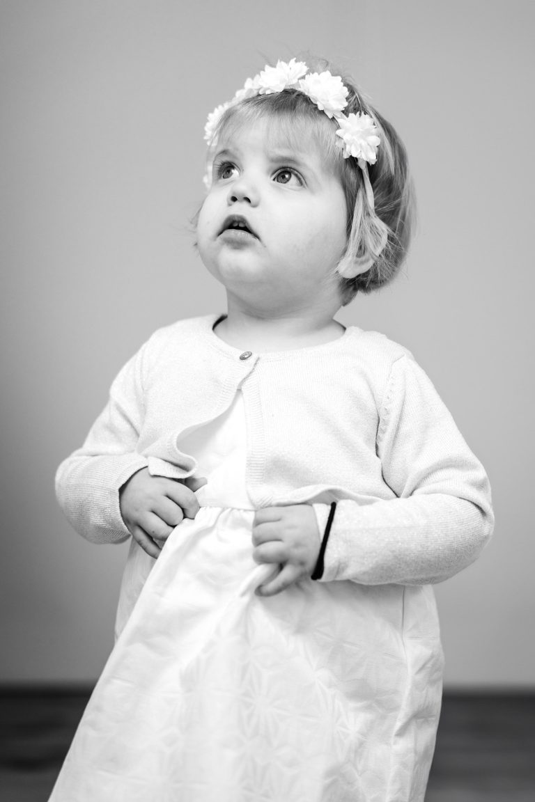 Une jeune enfant aux cheveux courts et clairs porte un bandeau fleuri et une robe de couleur claire avec un gilet. Elle regarde légèrement vers le haut avec une expression pensive. La photo est en noir et blanc, avec un fond uni et un éclairage doux.