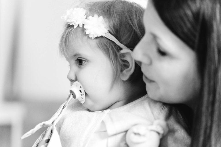 Une femme tient affectueusement un jeune enfant portant un bandeau de fleurs et une tétine accrochée à ses vêtements. L'enfant regarde au loin tandis que la femme sourit doucement, son visage près de celui de l'enfant. La photo est en noir et blanc.