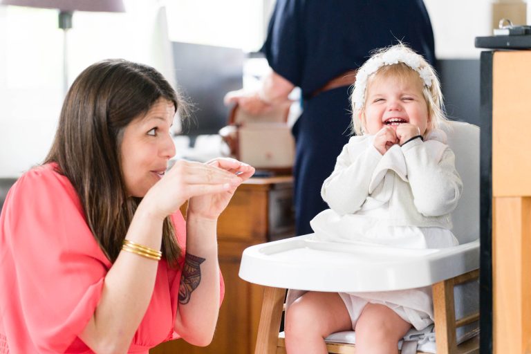 Une femme vêtue d'un haut corail fait des gestes ludiques avec ses mains pour amuser un bambin souriant dans une chaise haute. L'enfant, habillé en blanc avec un bandeau, rit joyeusement. La scène semble se dérouler à l'intérieur, un autre adulte étant flou à l'arrière-plan.