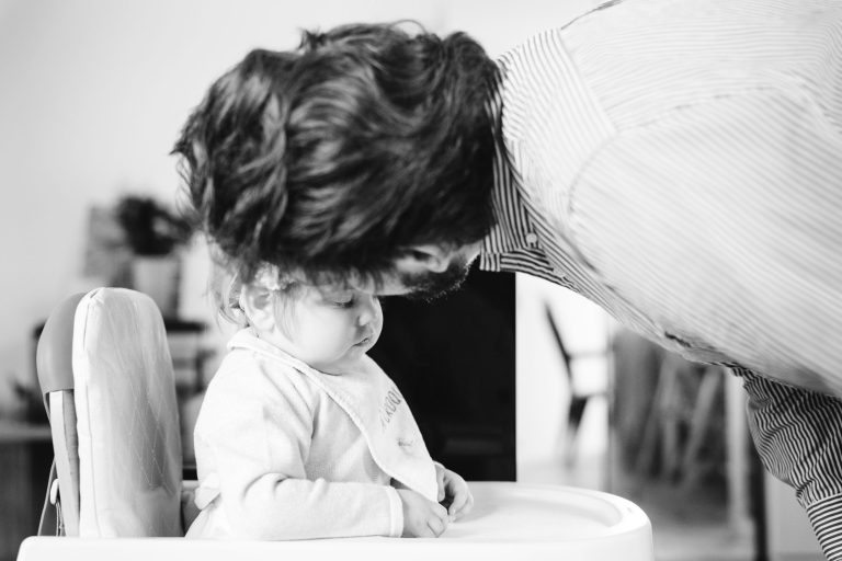 Un homme aux cheveux noirs se penche près d'un bébé assis dans une chaise haute. Le bébé, qui porte un bavoir et des vêtements clairs, regarde vers le bas. La scène est en noir et blanc, créant une atmosphère douce et intime. L'arrière-plan est flou.