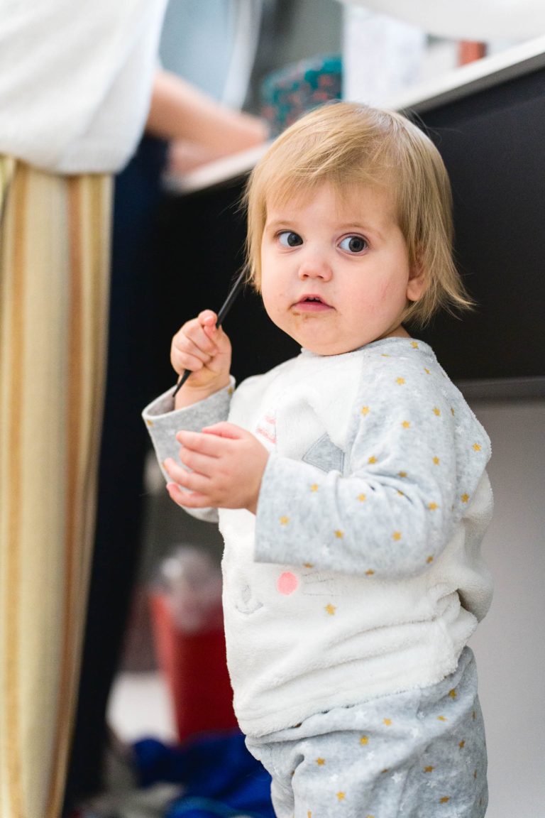 Un enfant en bas âge aux cheveux châtain clair, portant un pyjama gris à pois jaunes, tient une cuillère et une tasse. L'enfant regarde légèrement sur le côté avec une expression curieuse. Un adulte se tient à l'arrière-plan, près d'un comptoir, partiellement hors du cadre.