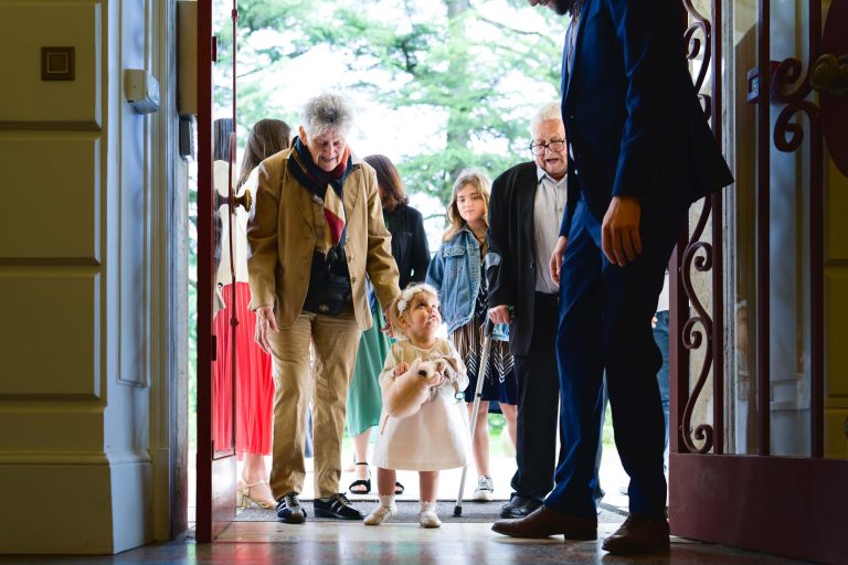 Une petite fille vêtue d'une robe blanche et d'une couronne de fleurs se tient dans l'embrasure d'une porte, tenant un ours en peluche, et regardant un homme en costume bleu. Plusieurs adultes et enfants, habillés pour un événement officiel, lui sourient et la suivent à l'intérieur par des portes ouvertes ornées. Des arbres sont visibles à l'extérieur.