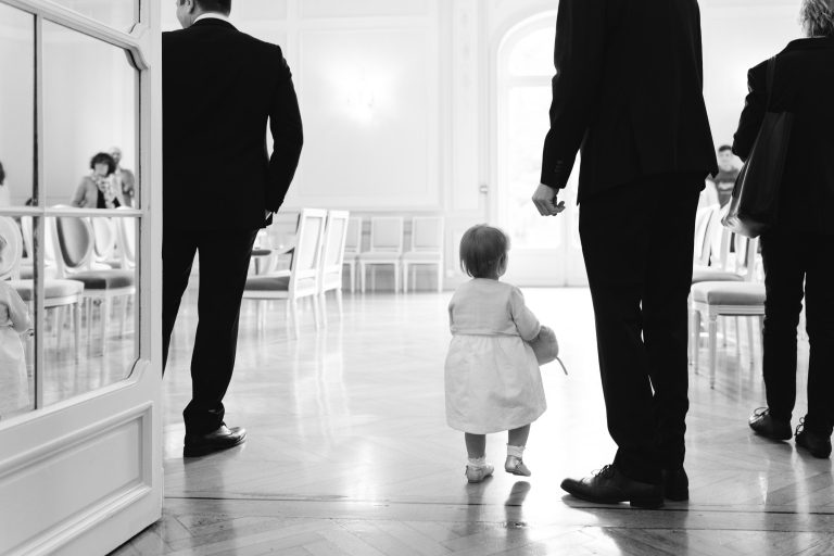 Un petit enfant vêtu d'une robe blanche marche sur un sol poli en tenant la main d'un adulte vêtu d'un vêtement sombre. D'autres adultes se tiennent à proximité, dans une pièce lumineuse et élégante, avec des rangées de chaises vides et de grandes fenêtres. La scène est en noir et blanc.