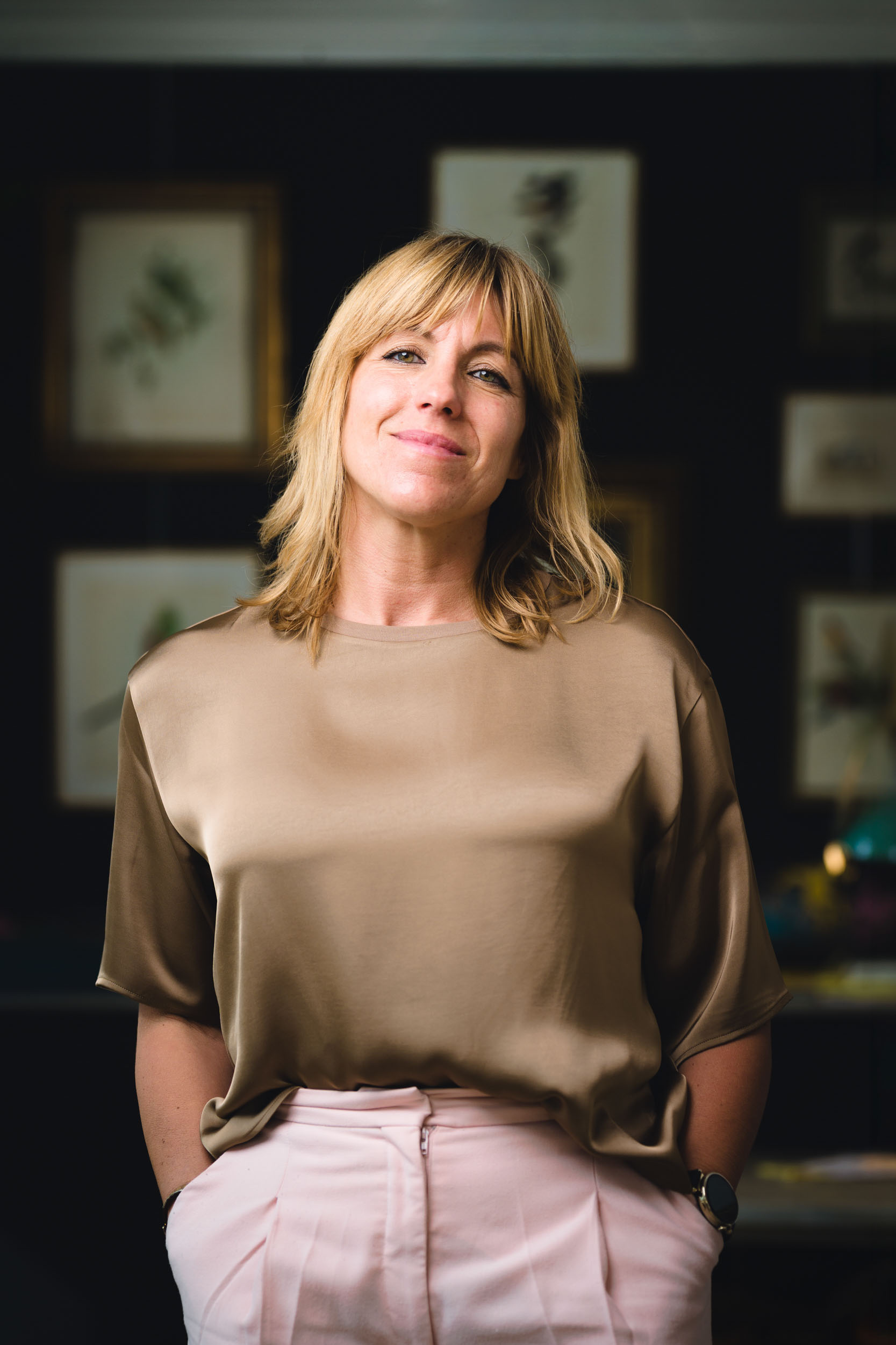 Une femme blonde pose pour un portrait corporate à bordeaux, derrière elle il y a un mur sombre avec des cadres photos. Elle est habillée avec un haut doré et un pantalon rose. Elle se tient debout et est appuyer sur un bureau ancien.